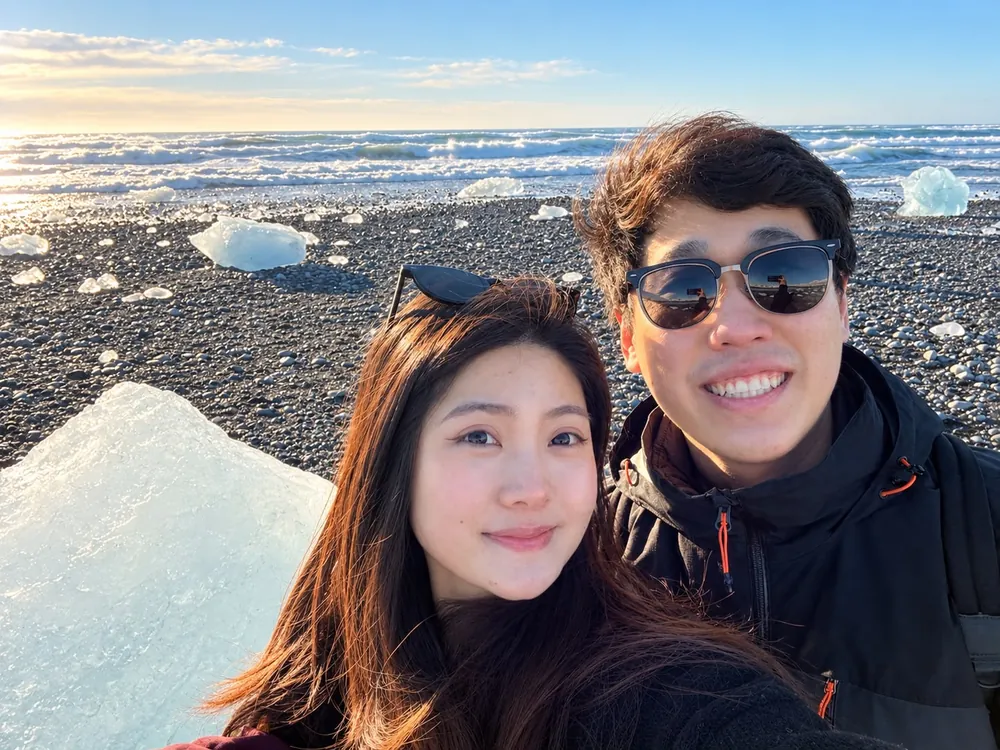 Camille smiling beside a companion on a pebble beach, with chunks of ice and the sea behind them